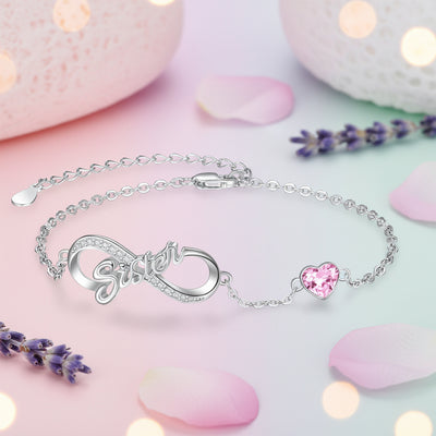 A silver chain bracelet featuring a central infinity symbol charm with the word 'Sister' in cursive, small clear rhinestones, and a pink heart-shaped crystal accent, shown on a white background.