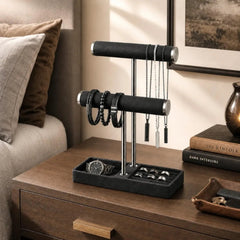 Black 2‑tier jewelry display stand on a bedside dresser holding necklaces, bracelets, rings, and a watch in a styled bedroom scene.