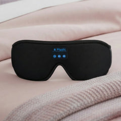 A black electronic muscle massager eye mask with a small blue-lit control panel, resting on a soft pink bed with a textured blanket in the foreground.