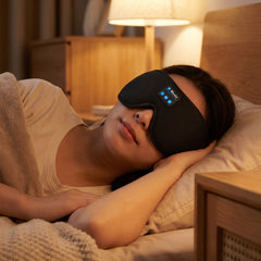 A woman lying in bed wearing a black sleep mask with blue LED indicator lights, in a warmly lit bedroom setting.