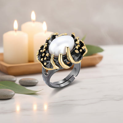 A silver-toned adjustable ring with a large white oval pearl center and textured black and gold floral-like detailing, shown on a marbel counter with cream candles in the background.