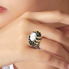 A large white oval baroque pearl in gold and black detailing, worn on a light skinned model hand near the face.