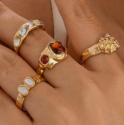 Four chunky gold rings with mixed gemstones shown on hand, featuring bold modern designs and polished gold-tone finish