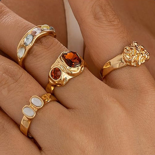 Four chunky gold rings with mixed gemstones shown on hand, featuring bold modern designs and polished gold-tone finish