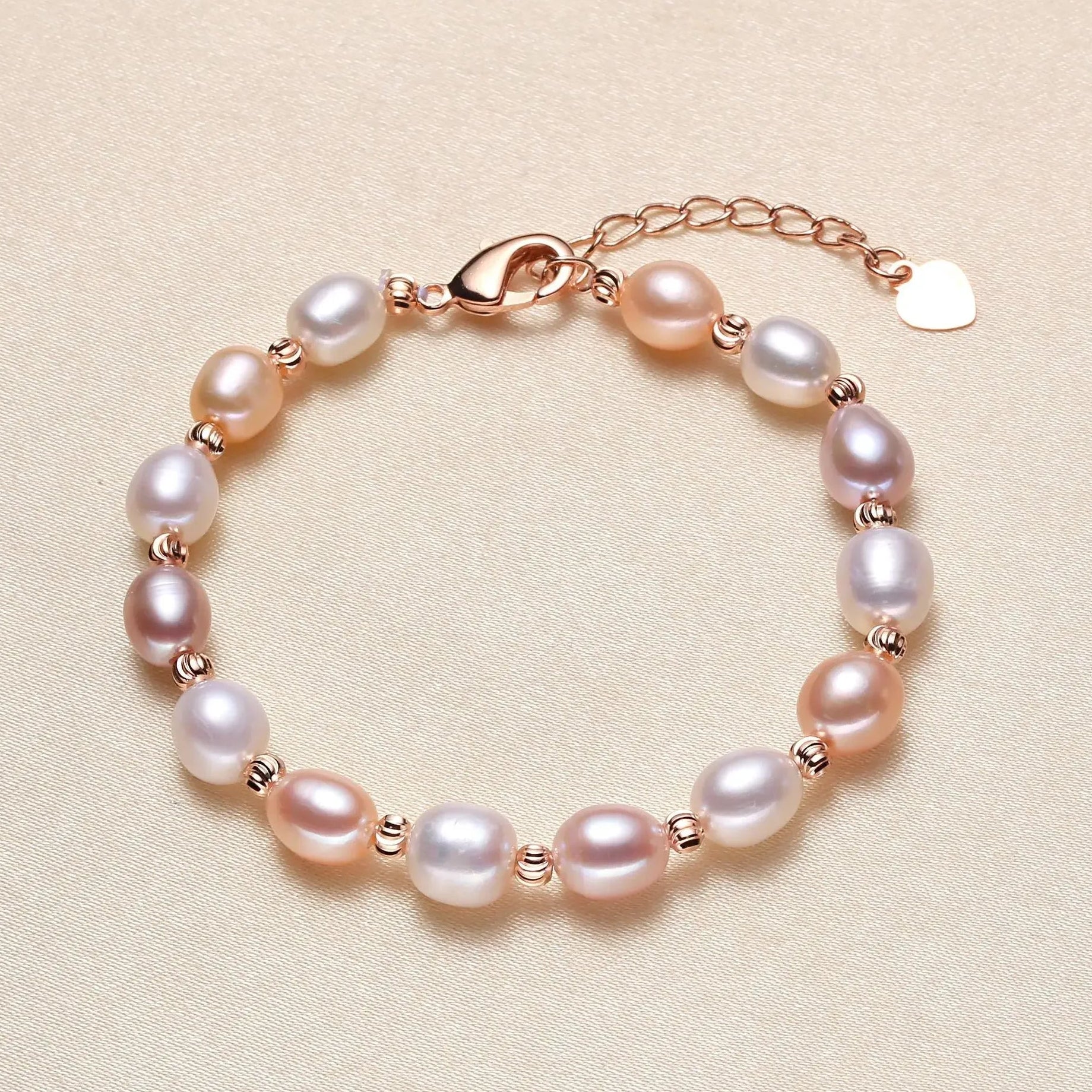 A freshwater pearl bracelet with pink, white, and purple pearls set in rose gold, featuring a lobster clasp, extender chain, and a small heart charm, displayed on a neutral beige background.