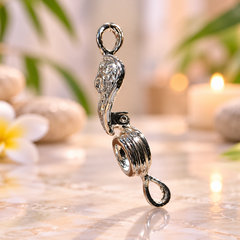 A polished silver-toned metal pendant charm with a stylized figure and loop details, standing upright on a reflective marble-like surface with a softly blurred spa-style background featuring a flower, stones, and a candle.