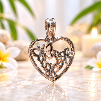 A polished silver heart-shaped pendant with intricate filigree butterfly and floral details, displayed on a reflective marble surface with white plumeria flowers and a soft-focus background of green leaves and a lit candle.