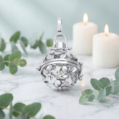 A silver-toned floral ball locket charm with a hinged lid and hanging loop, shown on a white table with candles and greenery in the background.