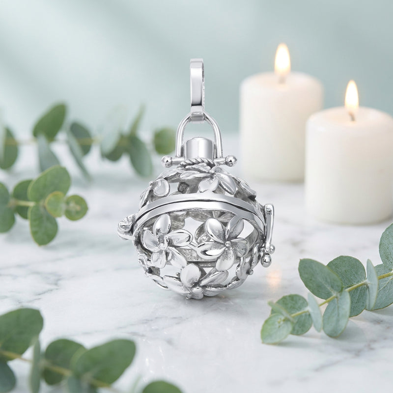 A silver-toned floral ball locket charm with a hinged lid and hanging loop, shown on a white table with candles and greenery in the background.