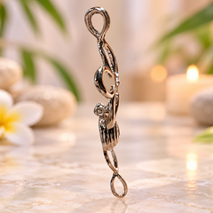 A polished silver-toned metal pendant shaped like a stylized infinity loop or spiral, standing upright on a light marble surface with a spa-like background featuring soft-focus stones, a white flower, and a lit candle.