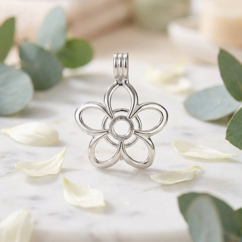 A close-up of a silver-toned metal flower-shaped charm or pendant on a white background with green leaves and white flower petals scattered about