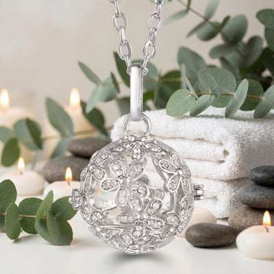 A silver filigree locket pendant necklace with a floral design, suspended from a matching silver link chain against a white towel with greenery and tan background.