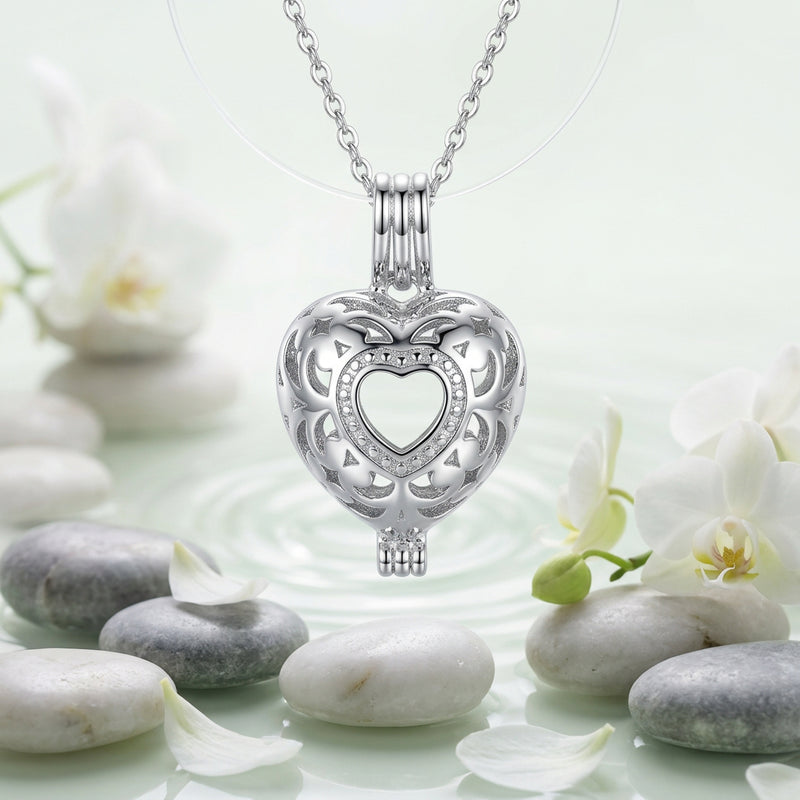 A close-up of a silver-toned metal heart-shaped charm or pendant on a white background with green leaves and white flower petals scattered about
