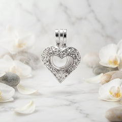 A close-up of a silver-toned metal heart-shaped charm or pendant on a white background with green leaves and white flower petals scattered about