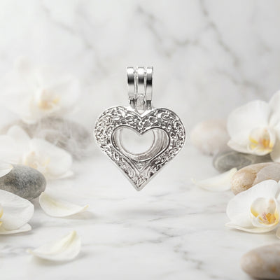 A close-up of a silver-toned metal heart-shaped charm or pendant on a white background with green leaves and white flower petals scattered about