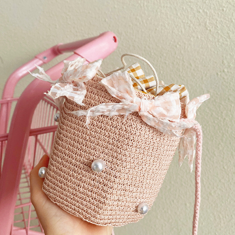A light pink woven straw bucket bag with pearl embellishments and ribbon bows, held in a hand in front of a pink shopping cart against a beige wall.