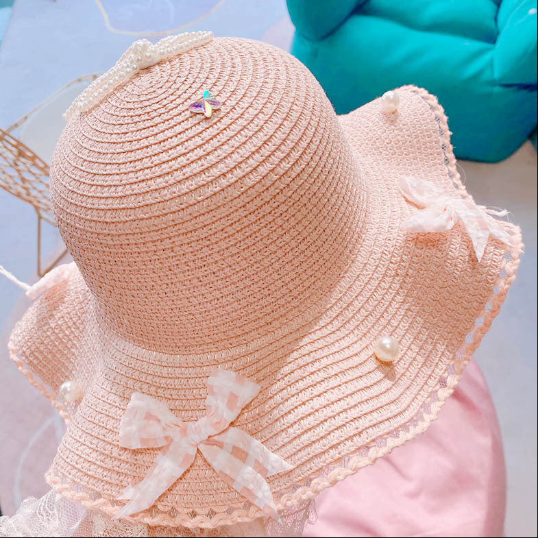 A light pink woven sun hat with a wide brim, decorated with white lace bows, pearl-like accents, and a small iridescent bow on top, shown against a soft pastel background.