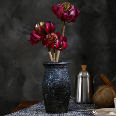 A dark blue speckled ceramic vase holding three artificial lotus flowers with deep burgundy petals and gold centers, styled on a table with a metal pitcher, yarn, and a cup against a dark textured backdrop.