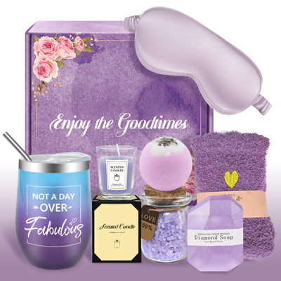 Purple-themed spa gift set with a satin sleep mask, gradient tumbler, scented candle, bath bomb, bath salts, fuzzy socks, and diamond-shaped soap arranged in front of a floral gift box.