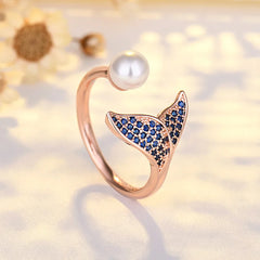 A rose gold open ring shaped like a fish tail, decorated with small blue stones, and topped with a white pearl, shown on a soft cream background with blurred flowers.