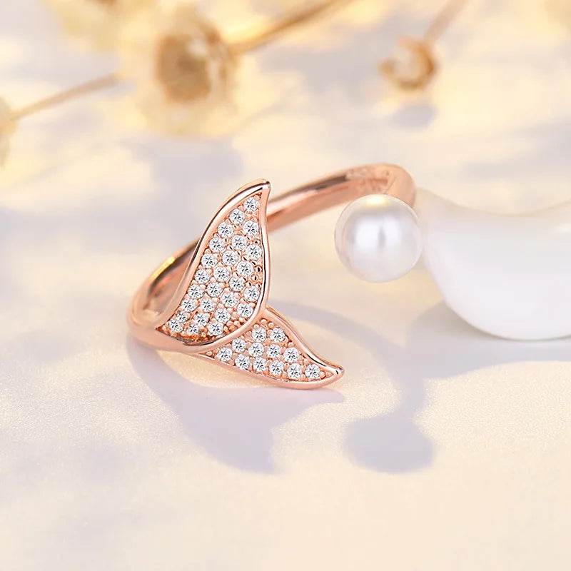 A rose gold open band ring featuring two curved, diamond-encrusted leaf-like shapes and a white pearl accent, displayed on a soft cream surface with blurred dried flowers in the background.