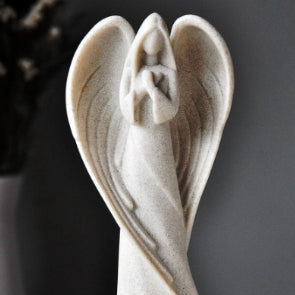 Angel sculpture with curved wings and clasped hands, made of light stone material, displayed against a soft gray background.