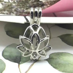 A silver-toned lotus flower pendant charm with a bail at the top, shown close up against a decorative background with green leaves and a pink object.