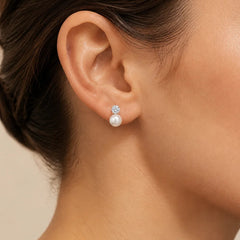 A close-up of a model's ear wearing a small stud earring with a clear gemstone and a white pearl drop.
