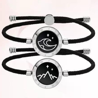 Two black braided bracelets with circular silver charms featuring wave and mountain designs on a light pink background.
