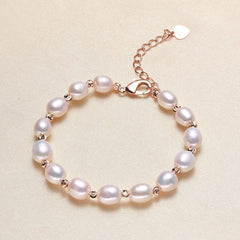 A white pearl bracelet with gold-toned metal spacers, clasp, and extender chain with a small heart-shaped tag, displayed on a textured beige background.