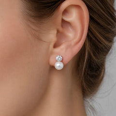A close-up of a woman's ear wearing a stud earring with a small clear crystal flower cluster on top of a white pearl drop.