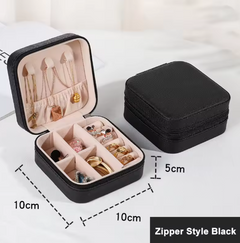 A black zippered jewelry box shown open and closed on a light fabric background. The open box reveals a light pink interior with compartments holding rings, necklaces, and earrings.