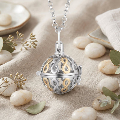 A silver and gold ball-shaped locket pendant necklace with an intricate filigree design, shown on a clean tan linen and stones background.