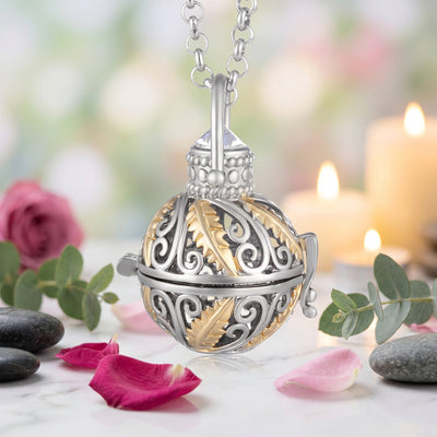 A silver and gold ball locket pendant necklace with intricate filigree scrollwork and leaf motifs, suspended from a silver link chain on a soft green and candle background.