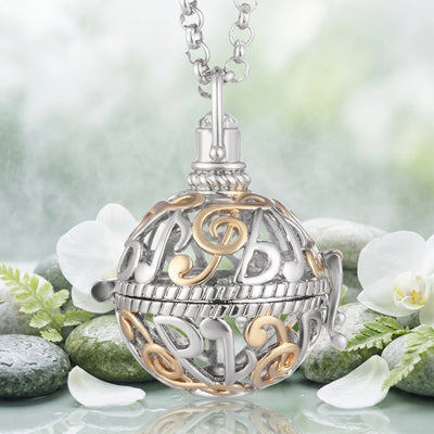 A silver and gold ball-shaped locket pendant necklace featuring musical notes and treble clefs, suspended from a silver link chain against a light green and white orchids background.
