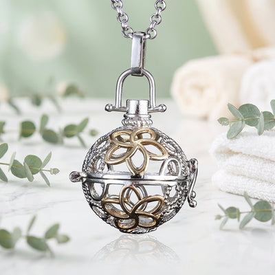 A silver and gold-tone ball-shaped locket pendant necklace with a floral design, shown on a light white and green background.