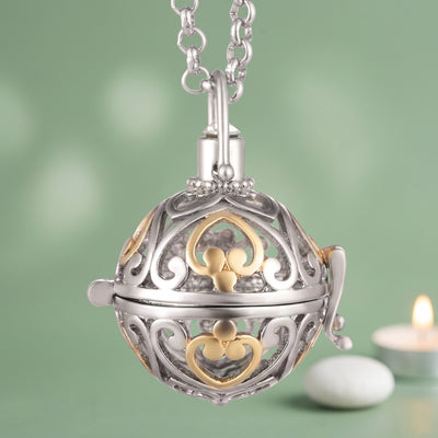 A silver and gold two-tone ball locket pendant necklace on a white background. The intricate filigree design features a silver heart and a gold clover motif, with a silver bail and chain on a green background
