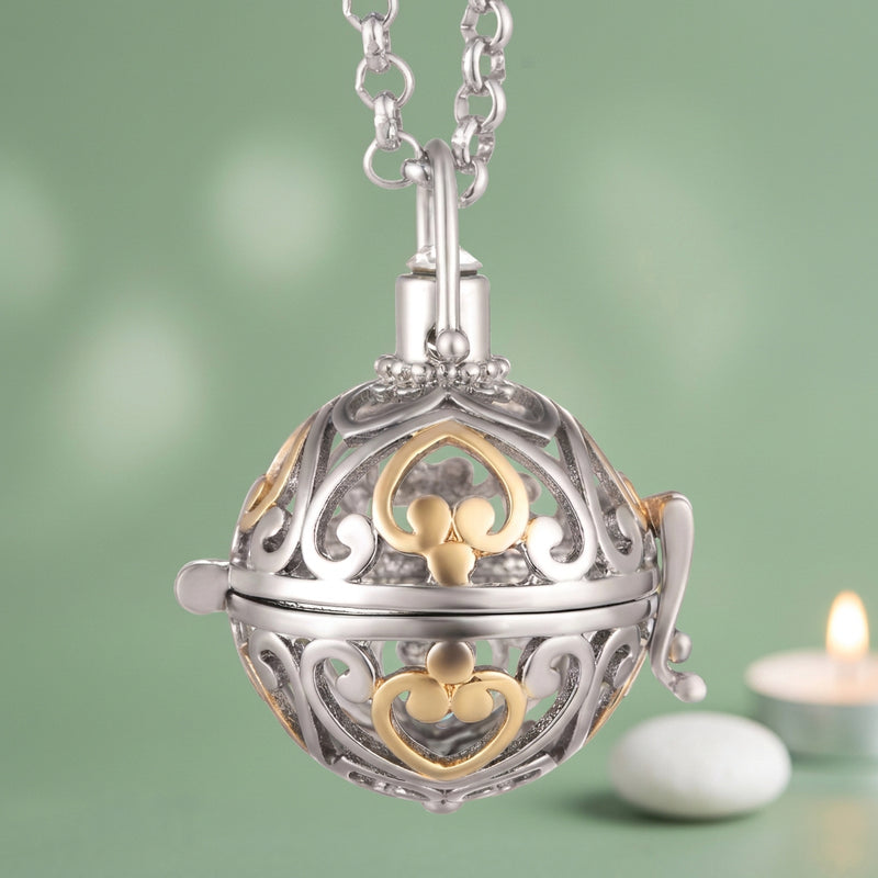 A silver and gold two-tone ball locket pendant necklace on a white background. The intricate filigree design features a silver heart and a gold clover motif, with a silver bail and chain on a green background