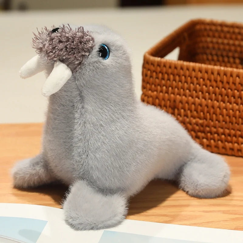 Gray walrus plush with white tusks, whiskers and blue-rimmed eyes sitting beside a wicker basket.
