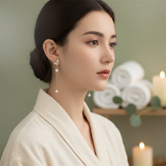 A close-up of a person wearing a dangling earring. The earring features a rhinestone-encrusted hearat-shaped top with a dangling chain and two white pearl-like beads.