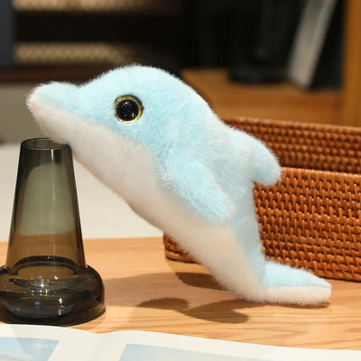 Light blue and white dolphin plush with a gold-rimmed black eye beside a dark glass and wicker basket.
