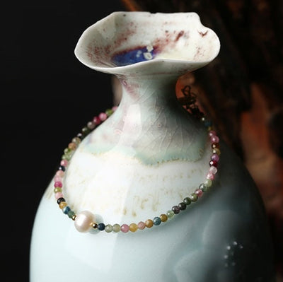 Delicate multicolor beaded bracelet with a single white pearl draped around a glossy blue‑green ceramic vase, shown against a dark background.
