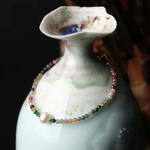 Delicate multicolor beaded bracelet with a single white pearl draped around a glossy blue‑green ceramic vase, shown against a dark background.