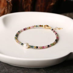 Multicolor beaded fashion bracelet featuring small round beads in pink, green, blue, and gold with a single white pearl accent, displayed on a textured stone surface.