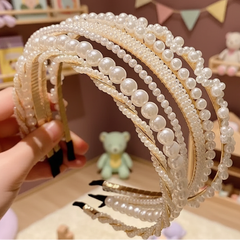 Hand holding multiple pearl-adorned headbands in assorted designs with gold accents, displayed in a soft pastel room with cozy decorative elements.