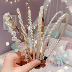 Hand holding five pearl-adorned headbands in assorted designs, featuring small and large pearls with gold-accented patterns, displayed in a soft pastel room with fairy lights.