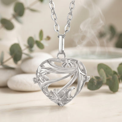 A silver heart-shaped locket pendant necklace with an intricate floral filigree design, suspended from a matching silver link chain against a tan and green plants background.