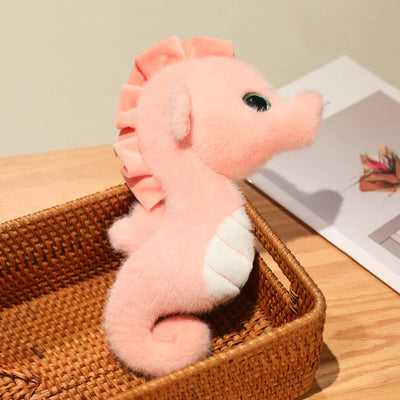 Pink seahorse plush with a white belly, curled tail and ruffled crest sitting in a wicker basket.