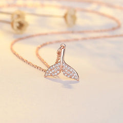 A rose gold mermaid tail pendant necklace adorned with small clear crystals, resting on a light beige surface with a matching chain and soft blurred decorative elements in the background.