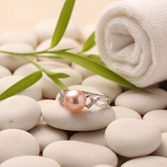 Pink freshwater pearl ring with a silver band and gemstone accents displayed on smooth white pebbles with bamboo leaves and a spa-style background.